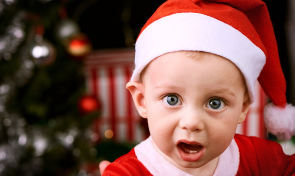 9 month old baby boy with shocked expression looking into the camera wearing a Santa Claus outfit for the christmas festive season.