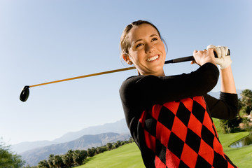 Happy young woman swinging club on golf course with mountains in background