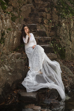 Young woman with blue eyes dressed bride sitting on a stone stairs in the countryside near a river. 