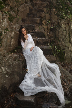 Young woman with blue eyes dressed bride sitting on a stone stairs in the countryside near a river. 