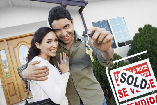Portrait of a happy couple holding keys in front of their house by sold sign