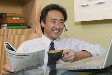 Happy businessman talking on headset while using laptop and reading newspaper