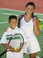 Portrait of happy mother and son with balls and racquet by the net on tennis court 