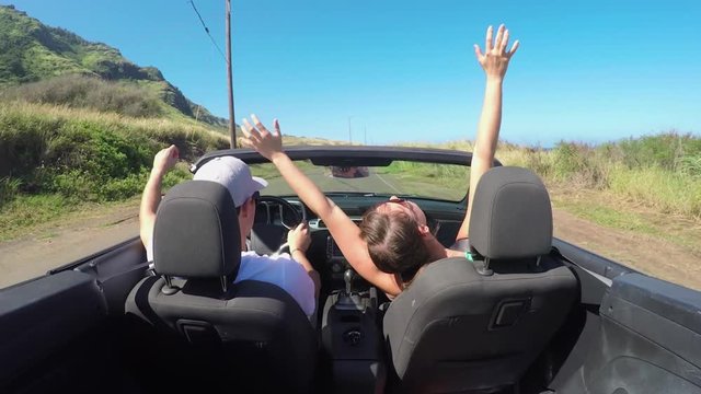 CLOSE UP: Happy couple with hands raised driving convertible along coastal road