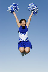 Full length of excited cheerleader with pompoms in midair against blue sky