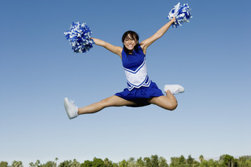 Full length of an excited young cheerleader jumping with pom-poms against sky