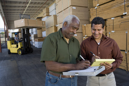 Happy storehouse workers writing on clipboard with man driving forktruck in the background