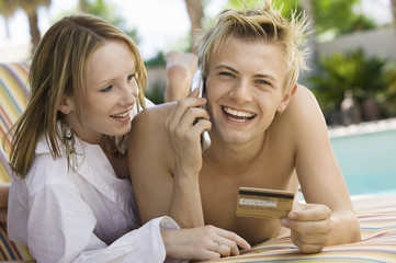 Young couple on deck chair by pool man making credit card purchase on mobile phone portrait