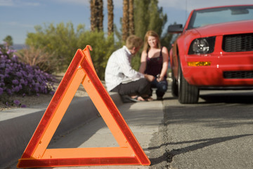 Emergency stop sign in foreground with blurred couple sitting by broken down car