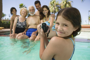 Fototapeta na wymiar Portrait of smiling girl with video camera recording family in swimming pool