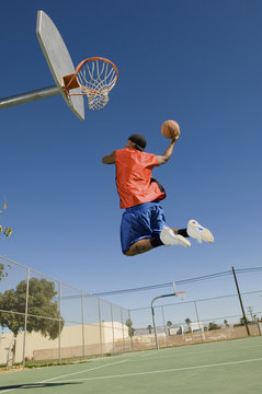 Low angle view of young man dunking basketball into hoop against clear blue sky