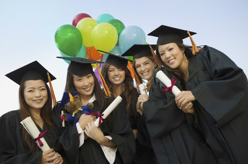 Portrait of happy female students with diplomas and balloons standing against sky