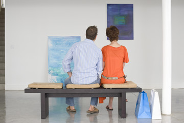 Rear view of couple looking at paintings while sitting on bench in art gallery - Powered by Adobe