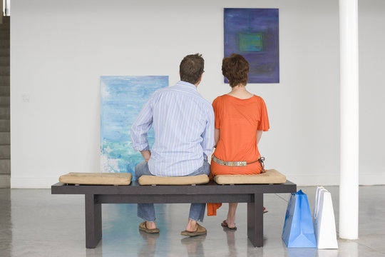 Rear view of couple looking at paintings while sitting on bench in art gallery