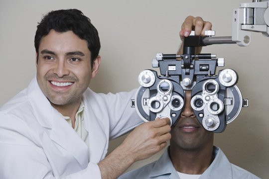 Happy male optometrist adjusting panels of phoropter while examining patient over grey background