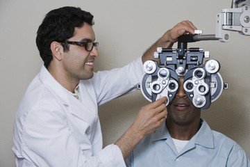 Mature male optometrist adjusting panels of phoropter while examining patient over grey background