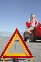 Closeup of warning triangle in front of a blurred woman on call by red car on an empty road