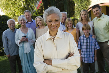 Portrait of happy senior woman standing with hands folded and family in background at garden