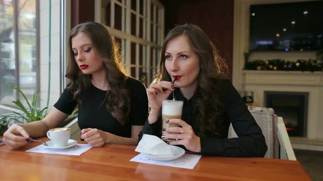 Two friends drinking coffee in a cafe. Sexy girl in black dresses drinking a latte. Two brunettes with red lipstick sitting in a restaurant.