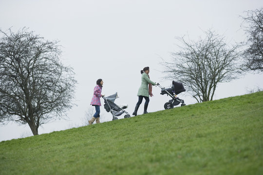 Full Length Of Young Mothers Pushing Baby Strollers Uphill In Park