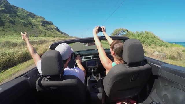 CLOSE UP: Happy couple driving in red convertible, young woman taking pictures 