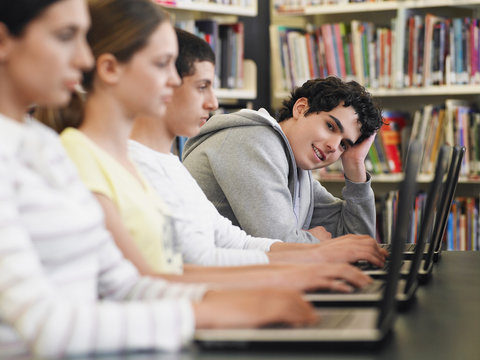 Portrait of teenage boy with friends in row using laptops in library