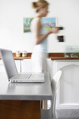 Young woman with coffeepot in kitchen walking by laptop in foreground