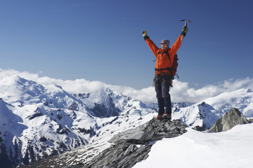 Male mountain climber raising hands with icepick on top of snowy peak