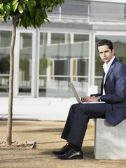 Portrait of young businessman using laptop on bench in plaza