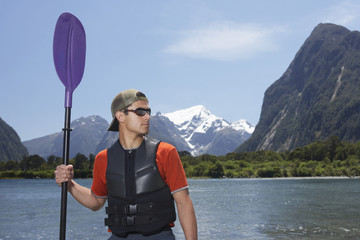 Young serious man holding an oar on shore of mountain lake