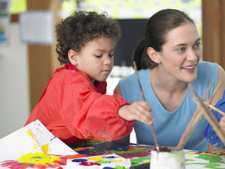 Young boy painting beside teacher in art class