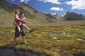 Obraz premium Man giving woman piggyback ride through pond near mountains