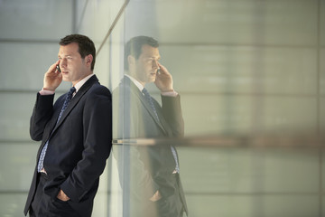 Middle aged businessman using mobile phone while leaning on glass wall in office