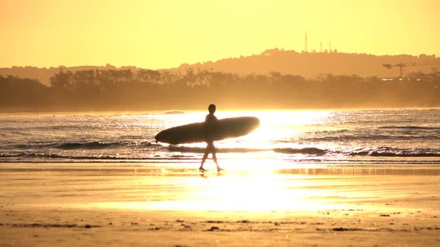 SLOW MOTION: Unrecognizable surfer girl carrying longboard surf at golden sunset