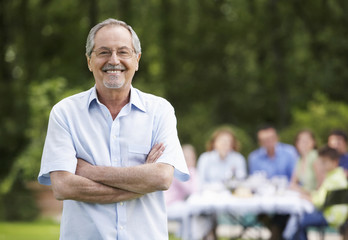 Portrait of senior man with arms crossed and family having lunch in background