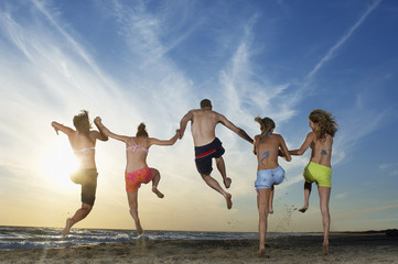 Full length rear view of young male and female friends jumping on sand while holding hands at beach