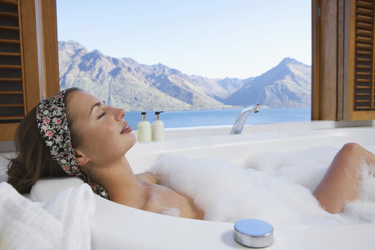 Side view of a young woman taking bubble bath with mountain lake outside window