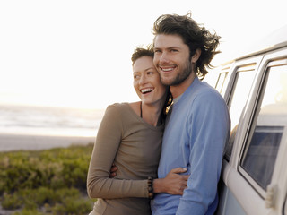 Smiling young couple embracing by campervan on beach - obrazy, fototapety, plakaty