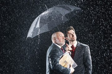 Young and middle aged businessmen watching rain from under umbrella