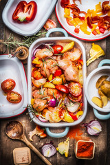 Tasty chicken with colorful vegetables and red sweet paprika in casserole, preparation on rustic wooden background and cutting cooking ingredients in enamelled bowls, top view