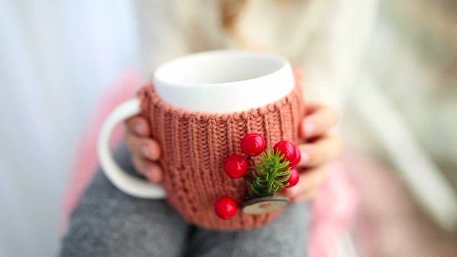 Close up of hands of child holding white mug in winter knitted cover festively decorated with Christmas details. Cute little girl sitting on windowsill, drinking hot tea. Real time full hd video.