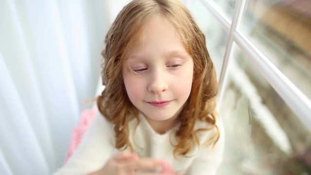 Portrait of cute happy little girl sitting alone on windowsill looking at camera. Child removing beautiful curly hair from face. Real time full hd video footage.