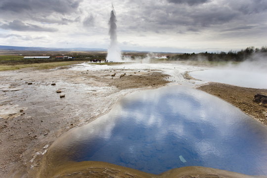 Blue water in geothermal pool with water spout from Strokkur Geysir visible in the distance, Geysir, near Reykjavik, Iceland