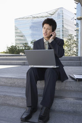 Young businessman using laptop and cellphone on outdoor steps