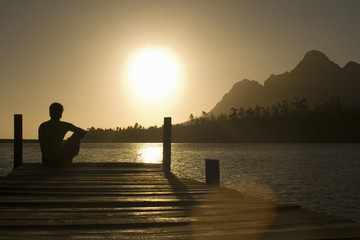 Rear view of man sitting on dock by lake enjoying sunset - Powered by Adobe