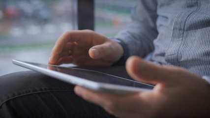 Young man in glasses sitting on a window sill in the hall while watching the news on the tablet.
