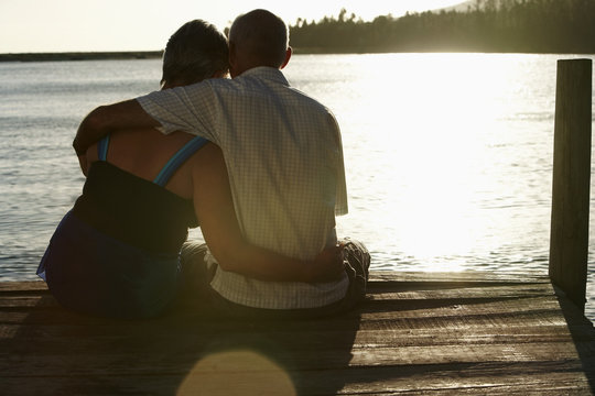 Rear View Of Happy Senior Couple Sitting On Edge Of Pier By Lake