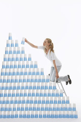 Side view of a young woman kneeling on ladder to stack plastic cups into pyramid against white background