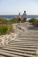 Rear view of boy and girl walking on boardwalk toward sea - obrazy, fototapety, plakaty