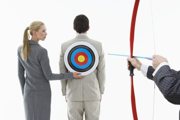 Business woman holding target to man's back while other man aims bow and arrow